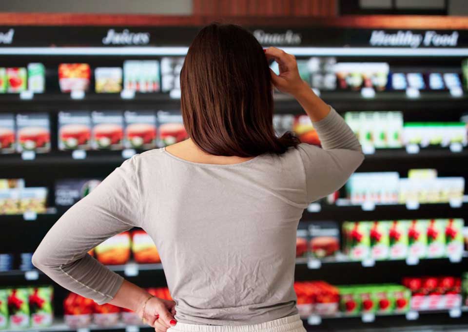 Eine Frau vor einem Warenregal in einem Supermarkt überlegt, welches Produkt sie kaufen soll.